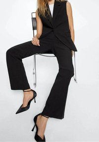 Pantalon noir sur mesure avec une veste sans manches, présentant un design structuré et une texture lisse, associé à des escarpins noirs à bout pointu.