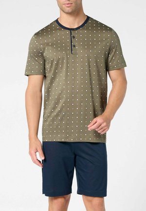 Uomo che indossa una maglietta henley a maniche corte verde oliva con piccolo motivo floreale e pantaloncini con coulisse blu navy, in piedi su sfondo bianco.