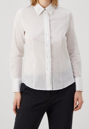 Kobieta ubrana w dopasowaną, białą koszulę na guziki z długim rękawem, z cienkimi pionowymi paskami, oraz czarne spodnie, stojąca na jednolitym tle.