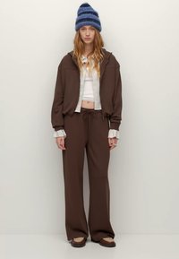 Brązowa bluza z kapturem z prążkowanymi ściągaczami i zamkiem błyskawicznym, zestawiona z pasującymi szerokimi spodniami. Dopełnieniem stylizacji jest niebiesko-szara, prążkowana dzianinowa czapka beanie.