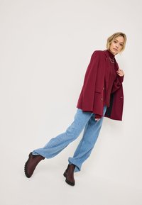 Blazer burdeos con un corte entallado y dos botones, combinado con un suéter de punto burdeos y jeans de pierna ancha azul claro. Botines marrones completan el look.