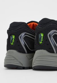 Černé sportovní boty s texturovaným síťovaným svrškem, šedou střední podrážkou a zelenými detaily. Mají výrazný podpatek a viditelnou výstelku.