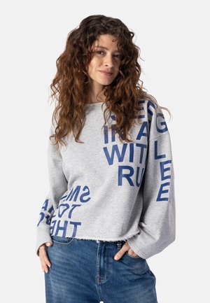 Giovane donna con capelli ricci che indossa una felpa grigia con grandi lettere blu e jeans blu, mani in tasca, sfondo neutro.