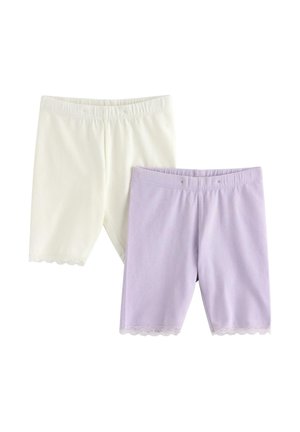 Ung pige står smilende iført en lilla, prikket kortærmet skjorte, hvide shorts med blonder, blomsterhat og beige flip-flops.