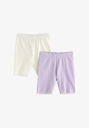 Ung pige står smilende iført en lilla, prikket kortærmet skjorte, hvide shorts med blonder, blomsterhat og beige flip-flops.