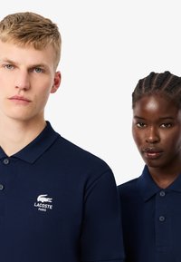 Granatowe koszulki polo z teksturowanej tkaniny. Posiadają małe białe logo Lacoste na lewej piersi, zapięcie na guziki oraz klasyczny kołnierzyk.