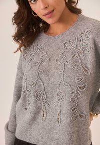 Suéter de lana gris con bordado floral y detalles recortados en el frente. Cuello redondo y mangas largas. Textura suave.