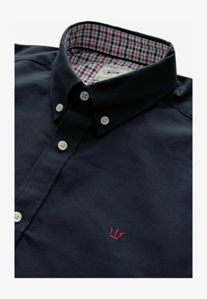 Navy-blaues Hemd mit Knöpfen und einem gemusterten Karomuster an der inneren Kragen. Verfügt über weiße Knöpfe und eine kleine rote Krone-Stickerei auf der Vorderseite.