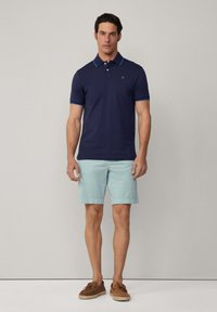 Marinblå polo skjorta med turkosa detaljer, korta ärmar och krage, parat med ljusblå shorts och bruna båt skor.