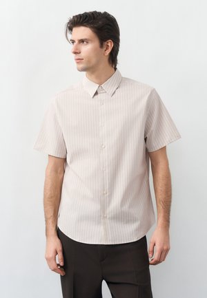 Junger Mann mit dunklem Haar, der ein hellbeiges Kurzarm-Hemd mit Knöpfen und schwarze Hose trägt, schaut nach links vor einem weißen Hintergrund.