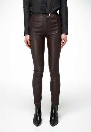 Frau trägt dunkelbraune, figurbetonte Kunstlederhose, schwarzes Hemd mit Knöpfen und schwarze, spitze Stiefeletten, steht vor einem weißen Hintergrund.