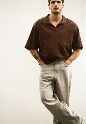 Mann mit Tätowierungen, der ein locker sitzendes dunkelbraunes Polohemd mit kurzen Ärmeln und hellgraue Jeans trägt, steht mit Händen in den Taschen und gekreuzten Beinen.