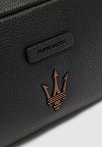 Zwarte, getextureerde leren pouch met een bruin metalen logo met drietandontwerp en een zwart etiket. Eenvoudige, rechthoekige vorm.
