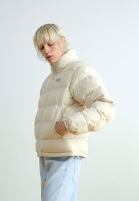 Beige puffer jacket with high collar, quilted design, and side pockets. Features logo on the chest and a smooth, shiny texture.