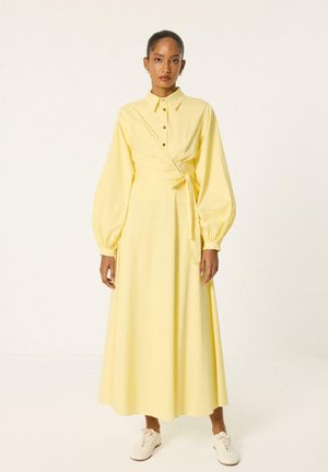 Femme portant une longue robe jaune à boutons avec des manches bouffantes et une ceinture à la taille, associée à des baskets blanches, debout devant un fond uni.