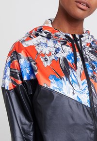 Barevná kapucínová bunda s květinovým potiskem v oranžové, modré a černé barvě, s lesklou texturou a zipovým zapínáním na přední straně.