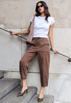 Mujer con camiseta blanca, pantalones marrones de pierna ancha y tacones, está de pie en las escaleras sosteniendo la barandilla con una mano y lleva un bolso beige al hombro.