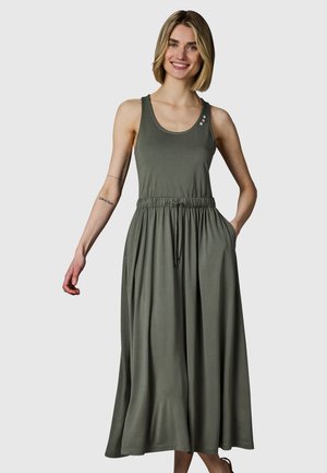 Smilende kvinde med kort blondt hår iført en ærmeløs olivengrøn maxikjole med elastisk talje og lommer, stående mod en ensfarvet baggrund.