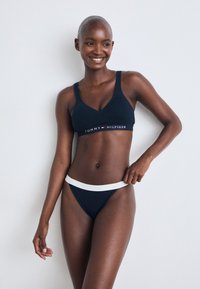 Temno modra spodnja perila, vključujoča bralette z V-izrezom in tangice z belim elastičnim pasom z logotipom "HILFIGER"; gladka tekstura tkanine.