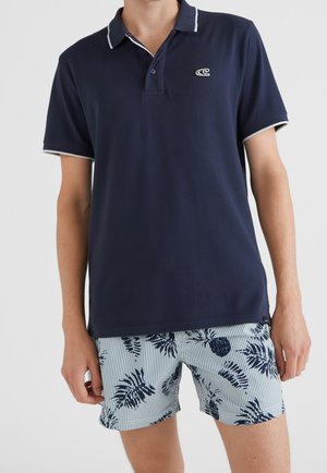 Marineblaues Polohemd mit kontrastierendem weißem Kragen, kombiniert mit hellblauen Shorts mit einem Muster aus Palmenblättern und Ananas. Einfarbige Stoffstruktur.