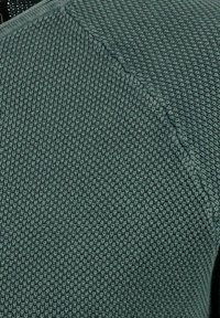 Strukturierter grüner Strickstoff mit einem subtilen Muster und sichtbaren Nähdetails. Er zeichnet sich durch ein weiches, gewebtes Design und einen leichten Glanz aus.