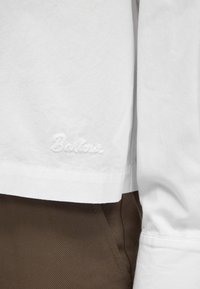 Chemise blanche à manches longues avec "Barker" brodé près de l'ourlet, associée à un pantalon marron.