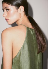 Green silk dress with a deep back neckline and thin straps, featuring a smooth texture and a flowing design. Hair styled in a sleek ponytail.