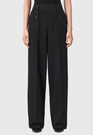 Højttaljede sorte bukser med vidde i benene og knapdetaljer ved taljen, båret med sorte spidse sko mod en ensfarvet baggrund.
