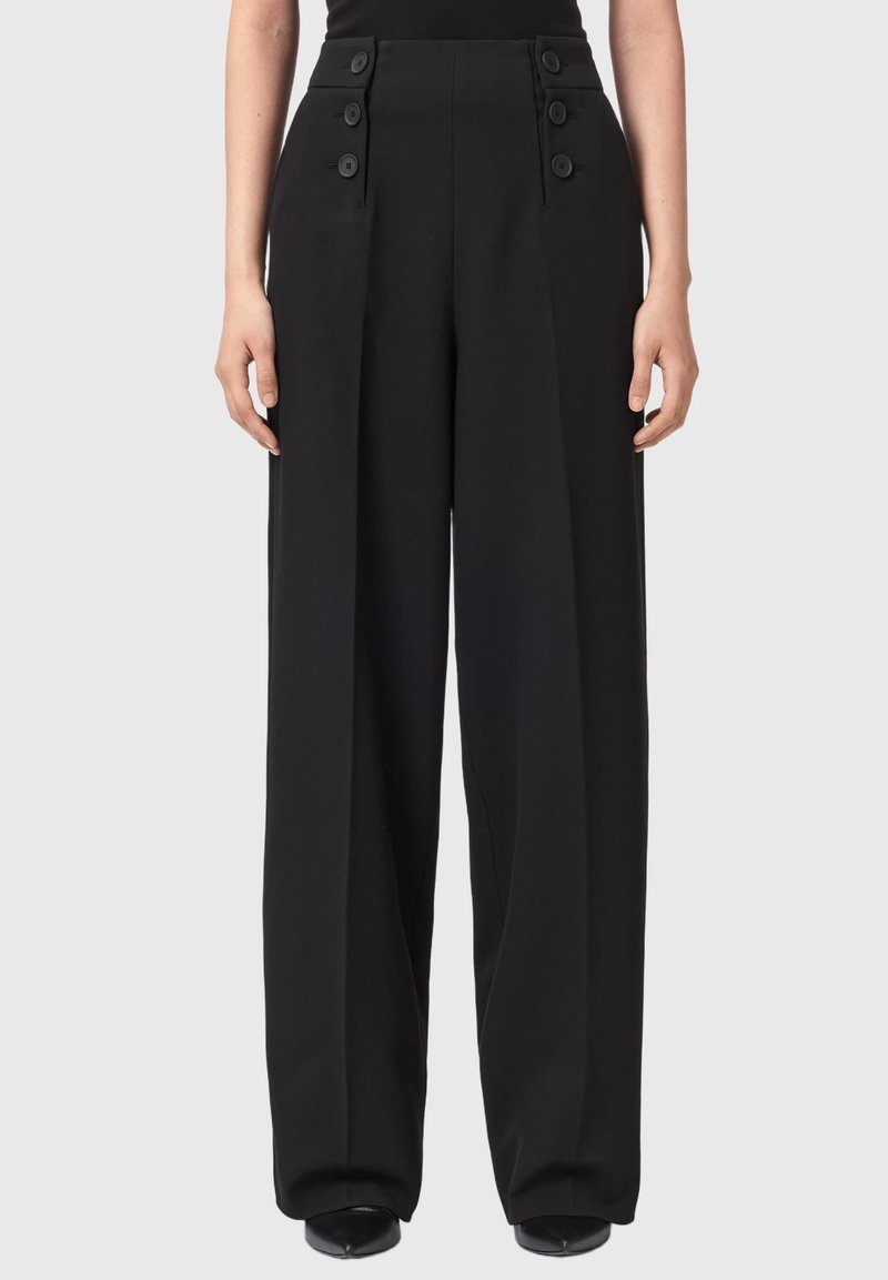 Schwarze, hoch sitzende, weit geschnittene Hosen mit Knopfdetails an der Taille, getragen mit schwarzen spitzen Schuhen vor einem schlichten Hintergrund.