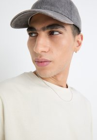 Casquette grise avec un bord courbé, t-shirt beige, collier en chaîne argentée, traits du visage mis en valeur, et arrière-plan neutre.