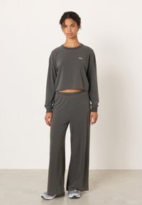 Jauna moteris stovi, dėvi tamsiai pilką ilgomis rankovėmis trumpą palaidinę ir derančias platėjančias kelnes bei pilkus sportbačius, paprastas fonas.