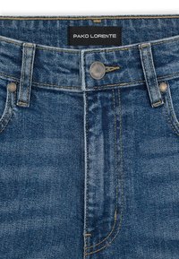 Granatowe dżinsy mają zapięcie na guzik, kontrastowe żółte przeszycia oraz metkę z napisem "PAKO LORENTE" przy pasku.