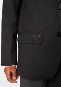 Suurendatud vaade mustast bleiserist, mille varrukas ja tasku klapp on kaunistatud peente pikseldatud ruutudega tikanditega, kandes tumedaid pükse ning käsi, mis asub ääre lähedal.