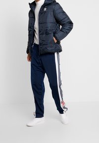 Padded check-pattern jacket in navy with a hood and zip closure, worn over a white shirt. Navy track pants with side stripes and buttons.