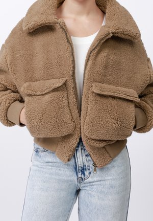 Person iført en lysebrun sherpa jakke med lynlås, store forlommer, ribstrikkede manchetter og kant, kombineret med lyseblå denimjeans.