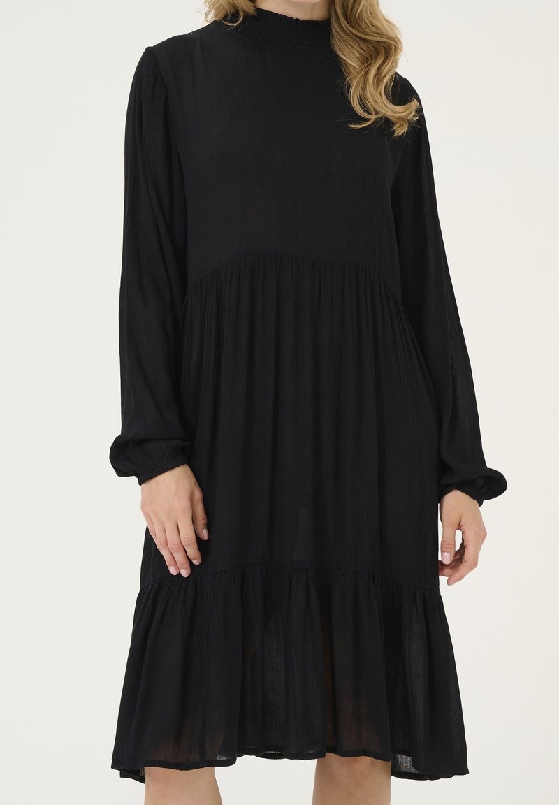 Femme portant une robe midi noire à manches longues avec une taille et un ourlet froncés, debout devant un fond clair uni.