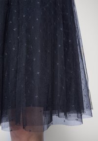 Marinblå tyllkjol med genomskinlig, lager-på-lager design, pryds av små, broderade stjärnmönster och har en mjuk, texturerad yta. Nederkanten är något fransig.