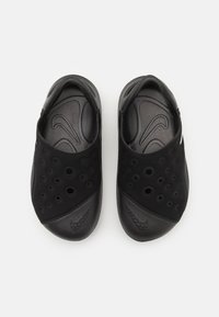 Black slip-on shoes featuring a textured upper with multiple circular cutouts, smooth rounded toe, and contrasting matte and glossy sections.