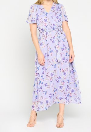 Woman wearing a light purple floral wrap dress with short flutter sleeves and tied waist, paired with beige strappy block heels.