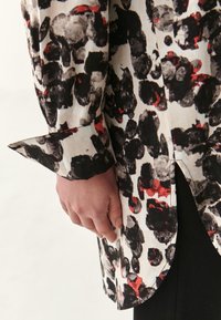 Blusa a maniche lunghe con base bianca e un audace motivo astratto nero, grigio e rosso. Il tessuto ha una texture liscia e una fessura laterale.