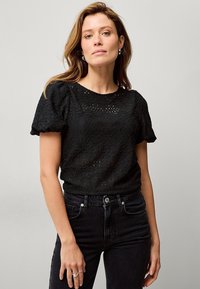 Schwarzes Spitzentop mit puffierten kurzen Ärmeln, rundem Ausschnitt und Cut-Out-Details. Kombiniert mit hochtaillierten schwarzen Jeans.