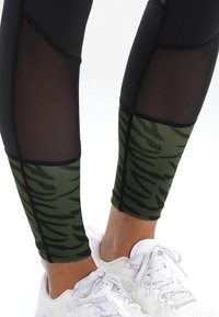 Czarne legginsy sportowe z zielonym wzorem w paski tygrysa na łydce, z przezroczystą siateczkową wstawką na górnej części nogi. Widać białe trampki.