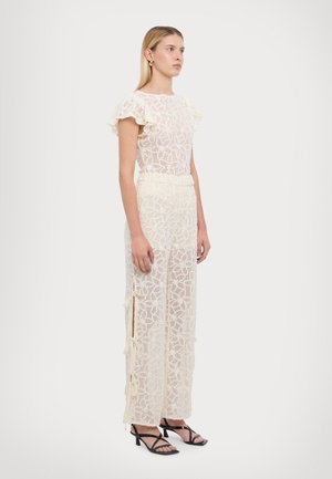 Cremfarbener Spitzen-Overall mit kurzen, rüschenbesetzten Ärmeln. Verfügt über ein florales Muster, eine gebundene Taille und seitliche Schlitze. Kombiniert mit schwarzen, schnürbaren High Heels.