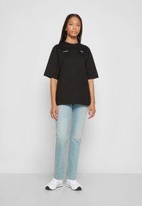 T-shirt de algodão preto com mangas curtas, apresentando uma marcação branca subtil na frente, combinada com jeans de denim azul claro e ténis brancos.