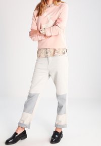 Sudadera rosa claro con diseño de renos blancos, superpuesta sobre un cuello con un patrón. Jeans de patchwork en blanco y gris. Zapatos negros.