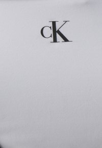 T-shirt en coton blanc avec une texture lisse et un logo "cK" noir. Le tissu présente un motif côtelé subtile.