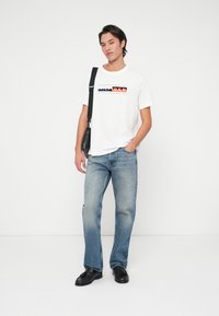 Young man wearing white graphic t-shirt, light blue jeans, black loafers, and shoulder bag, standing against plain white background.