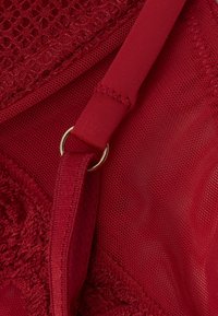 Rote Lingerie mit strukturiertem Netzdesign, glattem Band und einem goldfarbenen Metallschmuckdetail am verstellbaren Träger.