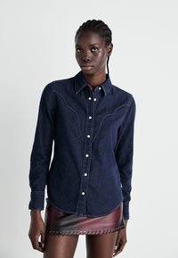 Granatowa denimowa koszula z zapięciem na guziki, długimi rękawami i dekoracyjnym haftem, w połączeniu z krótką burgundową skórzaną spódnicą z otworami.