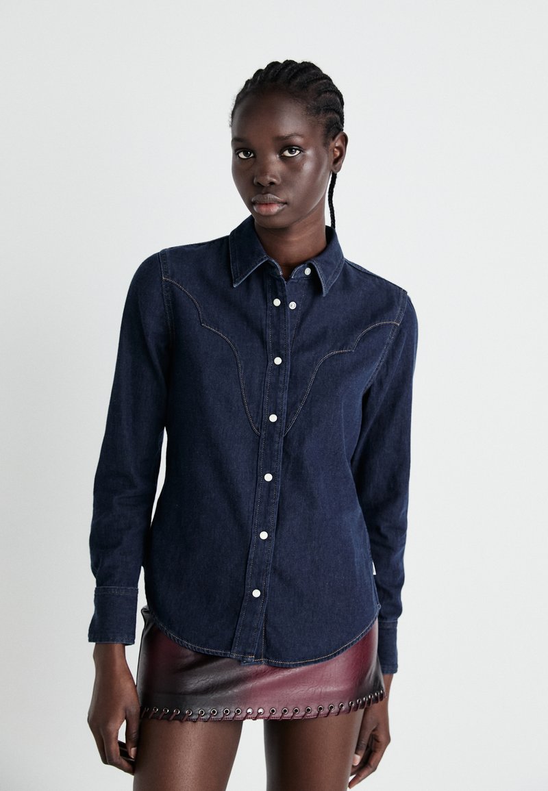 Granatowa denimowa koszula z zapięciem na guziki, długimi rękawami i dekoracyjnym haftem, w połączeniu z krótką burgundową skórzaną spódnicą z otworami.